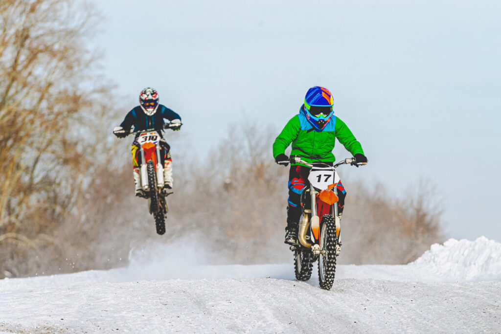Two dirtbike riders side by side on a snowy trail while wearing plenty of layers.